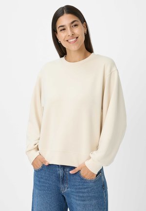 Crème-kleurige sweatshirt gemaakt van zachte stof, met een ronde hals, drop shoulders en geribde manchetten, gecombineerd met lichtblauwe spijkerbroeken.