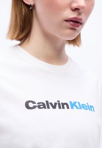 Κοντινό πλάνο ενός ατόμου που φοράει λευκό πουκάμισο με το λογότυπο "Calvin Klein" σε μαύρα και μπλε γράμματα στο στήθος.