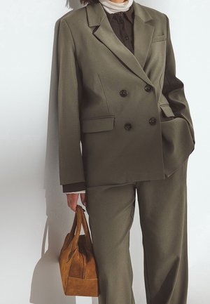 Personne portant un costume croisé vert olive, un col roulé blanc, une chemise noire, tenant un sac à main en daim marron avec poignées à côté d’elle.
