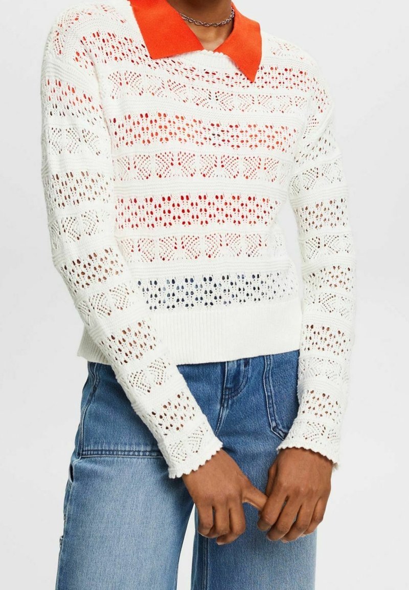 Weißer Strickpullover mit einem orangen Kragen. Verfügt über Lochmuster in wechselnden Streifen. Die Bündchen haben eine gewellte Kante. Getragen mit blauen Jeans.