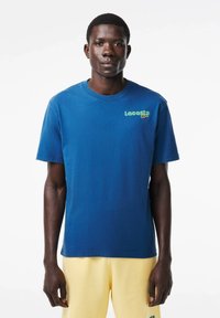 Camiseta de algodón azul con cuello redondo, que presenta un logo verde de Lacoste en el lado izquierdo del pecho y mangas cortas, combinada con pantalones amarillos.