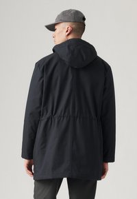 Zwarte capuchonjack met een tailleriem, lange mouwen en een gladde textuur. Draag het over grijze broek en combineer het met een grijze pet.