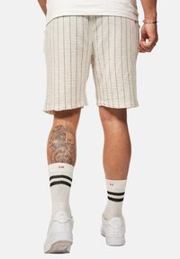 Helle beigefarbene Shorts mit schwarzen vertikalen Streifen, kombiniert mit weißen Socken mit schwarzen Streifen und weißen Sneakern. Auffälliges Tattoo am Bein.