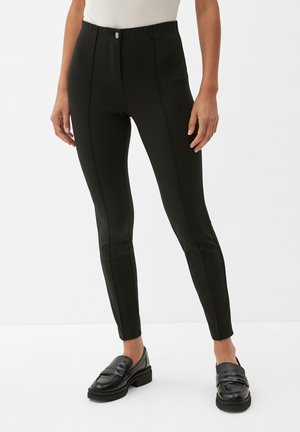 Pantalons noirs ajustés avec une taille haute, une fermeture éclair à l'avant et deux coutures latérales courbées. Le matériau a une texture lisse. Associés à des mocassins noirs.