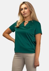 Zielona bluzka z krótkim rękawem, z dekoltem w kształcie łezki i złotymi akcentami. Gładki materiał, luźny krój, w komplecie z beżowymi spodniami.