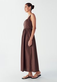 Rjava brezrokavna maksi obleka z oprijetim trupom, nabrano pasom in ohlapno krilo; nosi se z ravnimi črnimi sandali z zlatimi detajli.