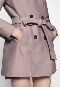 Femme portant un trench-coat rose poudré ceinturé avec de gros boutons noirs et les mains dans les poches latérales, montrée de la mi-cuisse à la taille.