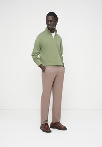 Πράσινο φούτερ με φερμουάρ, μπεζ παντελόνι και καφέ παπούτσια. Το μοντέλο στέκεται με τα χέρια στις τσέπες μπροστά σε απλό λευκό φόντο.