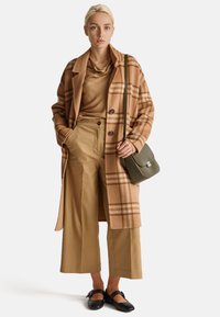 Brązowy w kratę wełniany płaszcz, beżowa bluzka z golfem, jasnobrązowe spodnie z szerokimi nogawkami i czarne płaskie buty. Zielona torebka Crossbody z fakturowanym detalem.