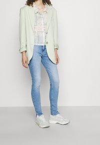 Blazer verde claro sobre una camisa de tonos pastel y estampado; combinado con jeans ajustados azul claro y zapatillas color menta. Texturas suaves y corte a medida.