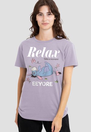 Woman wearing a lavender t-shirt with a relaxed cartoon Eeyore lying on his back, surrounded by flowers and the text "Relax Take a moment."