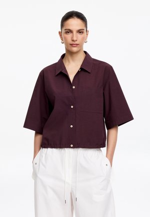 Femme debout face à l'avant, portant une chemise bordeaux foncé à manches courtes boutonnée et un pantalon blanc ample à cordon, les mains dans les poches.