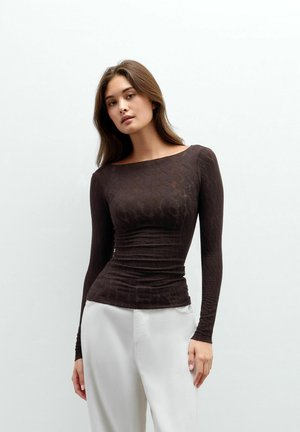 Joven mujer con cabello largo que lleva una camiseta de manga larga ajustada de color marrón oscuro con textura y pantalones blancos sueltos, de pie frente a un fondo liso.