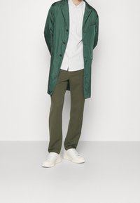 Vihreä satiinitakki valkoisen raidallisen paidan päällä, yhdistetty oliivinvihreisiin housuihin ja valkoisiin tennareihin. Takissa on kauluskäänne ja napit.