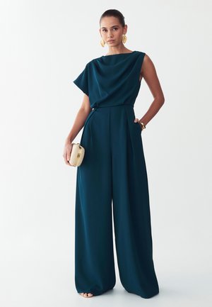 Donna che indossa una tuta color teal scuro a gamba larga con top drappeggiato, tiene in mano una piccola pochette beige ed è accessoriata con grandi orecchini e bracciali dorati.
