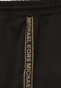 Černé tepláky s barevnou zlatou páskou se značkou po bocích. Měkká látka s elastickým pasem, jednoduchý design, žádné další vzory.