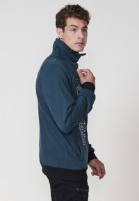 Chaqueta azul con estampado, con cremallera y un cuello alto, puños acanalados y un dobladillo negro en contraste. Presenta un texto gráfico llamativo en la parte delantera.