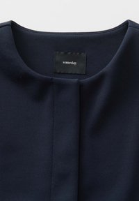 Navyblaue Bluse aus strukturiertem Stoff, runder Halsausschnitt und eine mittige Falte. Verfügt über ein schwarzes Etikett mit weißem Text, das am Hals angenäht ist.