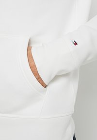 Bílá mikina s kapucí z měkkého materiálu, s klokankou kapsou, žebrovanými manžetami a malým logem na rukávu.