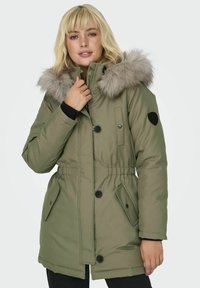 Parka verte olive avec capuche en fausse fourrure, boutons sur le devant et taille ajustable. Elle possède deux poches latérales et des poignets élastiques.