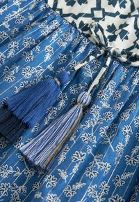 Tissu bleu avec des motifs floraux blancs et deux pompons bleus attachés à un cordon décoratif sur un vêtement.