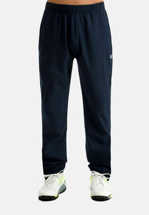 Homme portant un pantalon de sport noir à taille élastique et des baskets blanches avec des accents jaune néon, debout devant un fond uni.