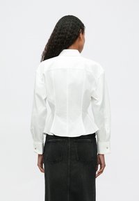 White peplum blouse with a structured back, featuring long sleeves and button cuffs, paired with a dark denim skirt. Smooth, crisp fabric.