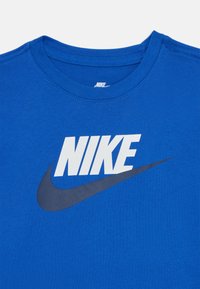 Plava pamučna majica s velikim bijelim "NIKE" logotipom i tamnoplavim swoosh dizajnom. Okrugli izrez s prošivenim rubom.