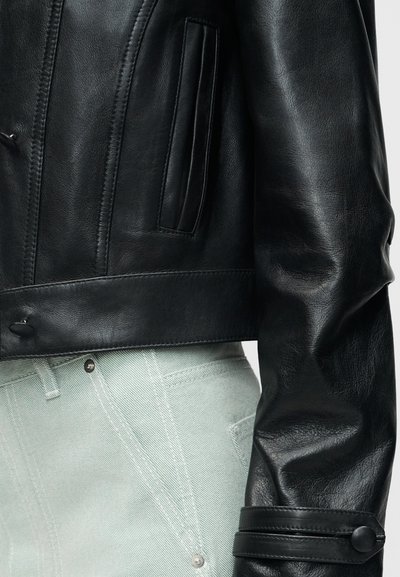 Κοντινό πλάνο μανικιού και τσέπης μαύρου δερμάτινου μπουφάν πάνω από ανοιχτόχρωμο τζην παντελόνι με ορατές λεπτομέρειες ραφής.