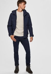 Dark blue puffer jacket with a hood, a light beige ribbed sweater, navy jeans, and brown hiking boots with black laces.