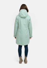 Mintgrøn lang frakke med hætte, med en lige pasform og knaplukkede ærmer. Bæres over mørke jeans og beige ankelstøvler.