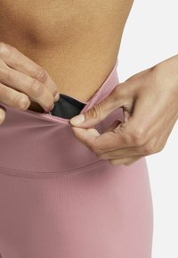 Ljusrosa sportleggings med en mjuk textur, med en svart säker fickstängning vid midjan som justeras av en hand.