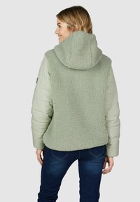 Veste vert clair avec un dos texturé et duveteux, des manches rembourrées et une capuche. Présente un devant lisse et une taille cintrée, assortie à un jean sombre.