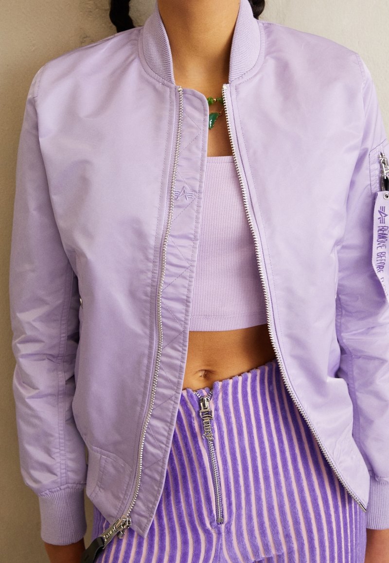 Fialová bomber bunda s žebrovaným límcem a manžetami, se stříbrným zipem a bočními kapsami. Pod ní se nachází odpovídající zkrácený top. Kalhoty s vertikálními pruhy.