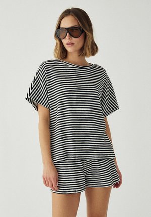 Kvinde iført store sorte solbriller og et matchende sort-hvidt stribet sæt med kortærmet top og shorts mod en neutral baggrund.