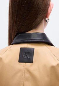 Καφέ μπουφάν με μαύρο δερμάτινο γιακά και τετράγωνο έμβλημα με το λογότυπο "cK". Ομαλή υφή, ραμμένο σχέδιο και εμφανείς λεπ τομέρειες ραφών.