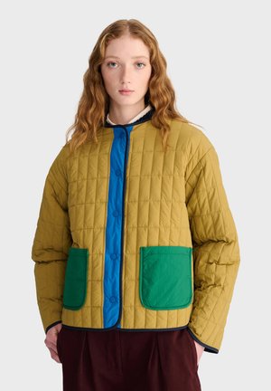 Jeune femme aux cheveux roux ondulés porte une veste matelassée moutarde avec une doublure bleue et des poches vertes à l'avant, debout devant un fond uni.