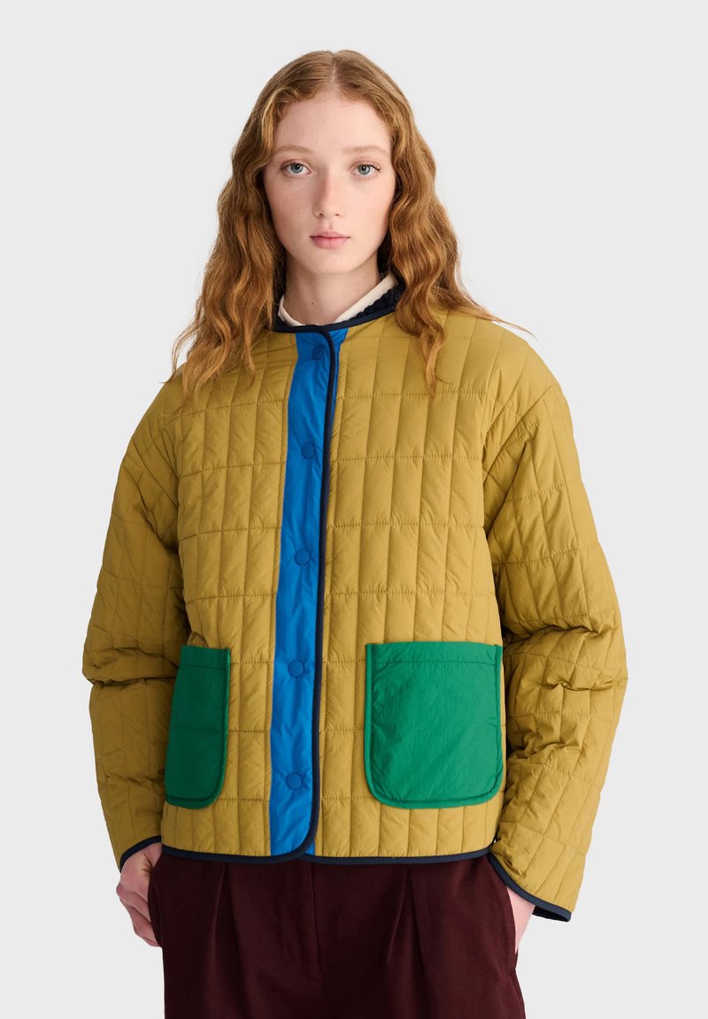 Jeune femme aux cheveux roux ondulés porte une veste matelassée moutarde avec une doublure bleue et des poches vertes à l'avant, debout devant un fond uni.