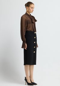 Braune transparente Bluse mit Schleifchen-Detail, kombiniert mit einem schwarzen Bleistiftrock mit goldenen Knopfakzenten. Schwarze High Heels runden den Look ab.