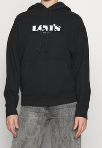 Svart huva sweatshirt med framficka och vit "LEVI'S" logotyp. Har dragsnören och ribbad mudd. Bärs med ljusgrå jeans.