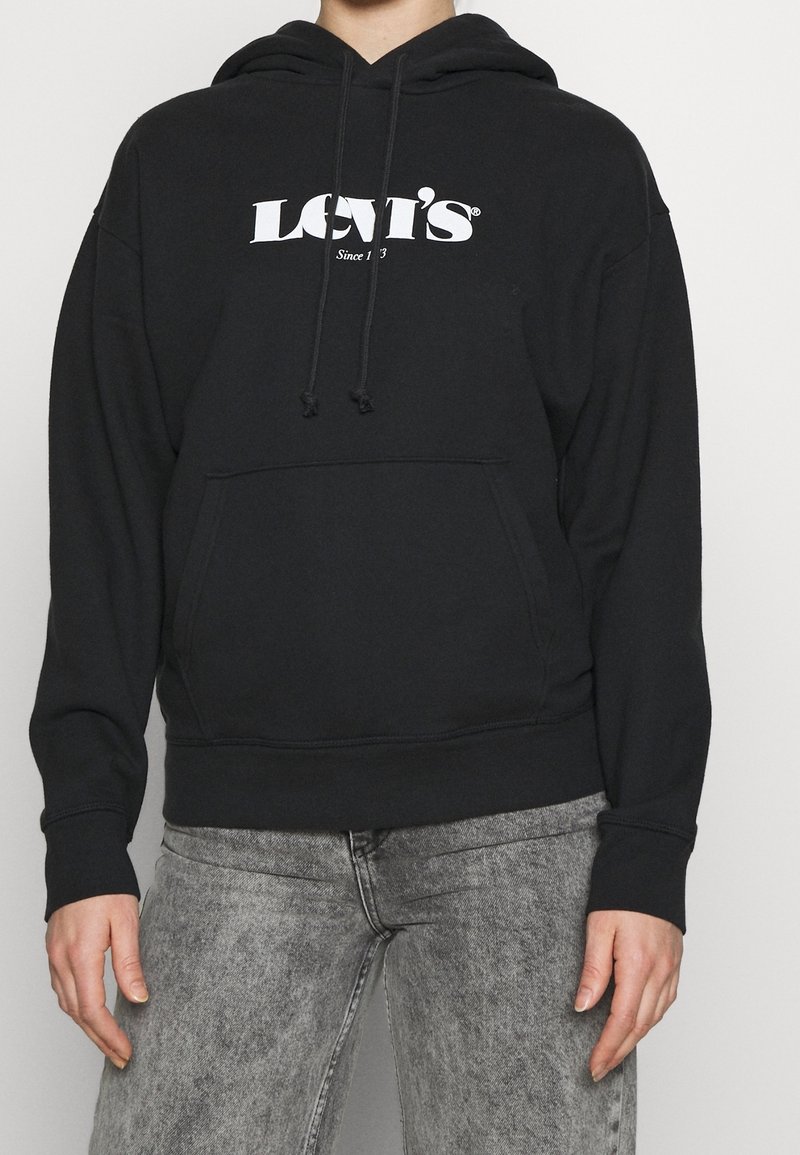 Svart huva sweatshirt med framficka och vit "LEVI'S" logotyp. Har dragsnören och ribbad mudd. Bärs med ljusgrå jeans.