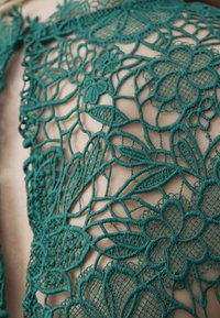 Superposition en dentelle turquoise avec motifs floraux et broderies complexes sur un fond neutre, mettant en valeur la texture et les éléments de design détaillés.