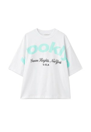 Giovane donna con capelli ricci che indossa una camicia bianca e pantaloncini verde menta, beve da un bicchiere riutilizzabile azzurro chiaro davanti a un cielo azzurro limpido.