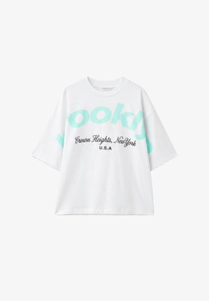 Junge Frau mit lockigem Haar, die ein weißes Shirt und mintgrüne Shorts trägt, trinkt aus einem hellblauen Mehrwegbecher vor klarem blauem Himmel.