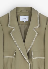 Blazer kaki cu cusături albe în zig-zag de-a lungul gulerului și buzunarelor, având rever cu tăietură și închidere cu un singur nasture.