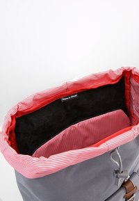 Harmaa reppu, jonka sisällä on punavalkoraidallinen vuori, mustasta pehmeästä kankaasta tehty pohja ja pieni sisätasku. Varustettu nyöri sulkimella ja metallipinnoilla.