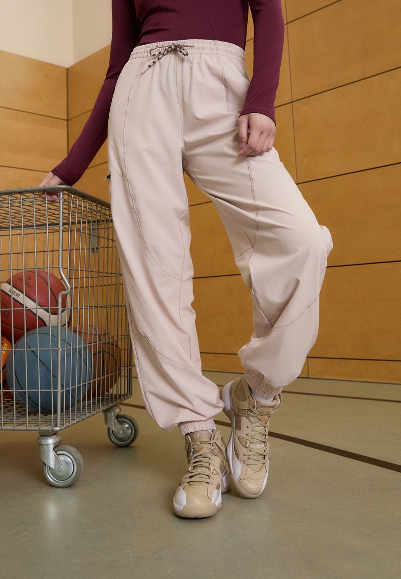 Lichtroze sportbroek met een drawstring taille en elastische boorden, gecombineerd met beige sneakers. Een draadmand met basketballetjes is in de buurt.