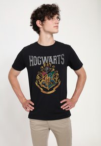 Černé bavlněné tričko s nápisem "HOGWARTS" ve šedém textu a barevný, detailní erb s zlatými akcenty pod nápisem.