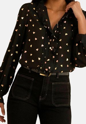 Kvinde iført en sort bluse med gyldne og lyserøde hjerteprint, kombineret med sorte højtaljede bukser og et sort bælte med en guldfarvet spænde.