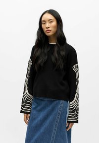 Svart genser med lange ermer og innviklede hvite linjemønstre; kombinert med et denimskjørt. Glatt tekstur og avslappet passform.